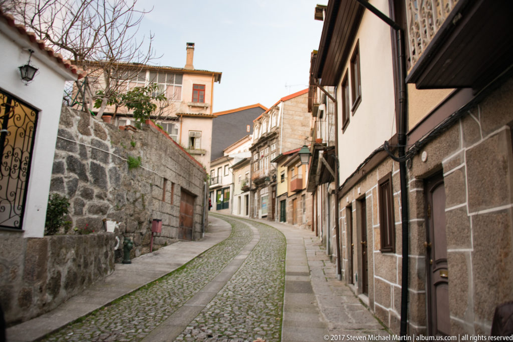 Narrow curving cobbled lane rising between granite walls and traditional houses with green trim, tiled roofs, and projecting upper stories.