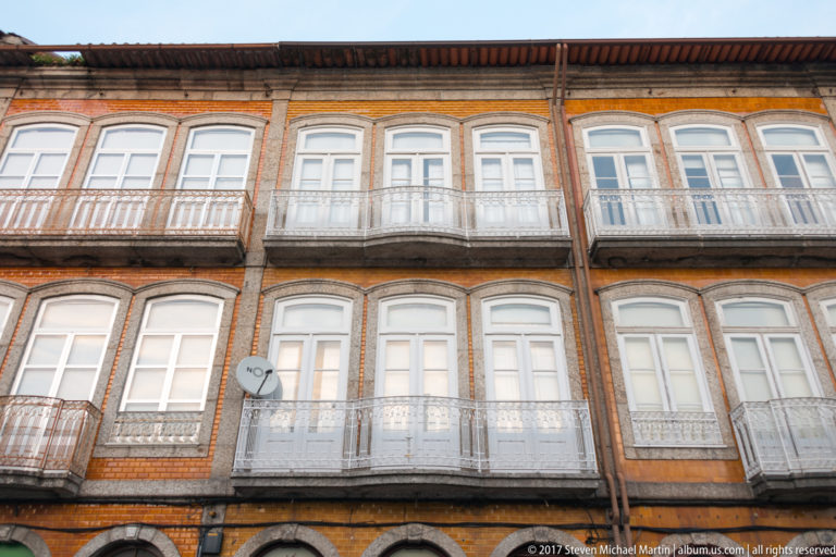 Upper stories of an orange historic façade with repeated white windows, slender iron balconies, granite bands, and dark projecting eave.