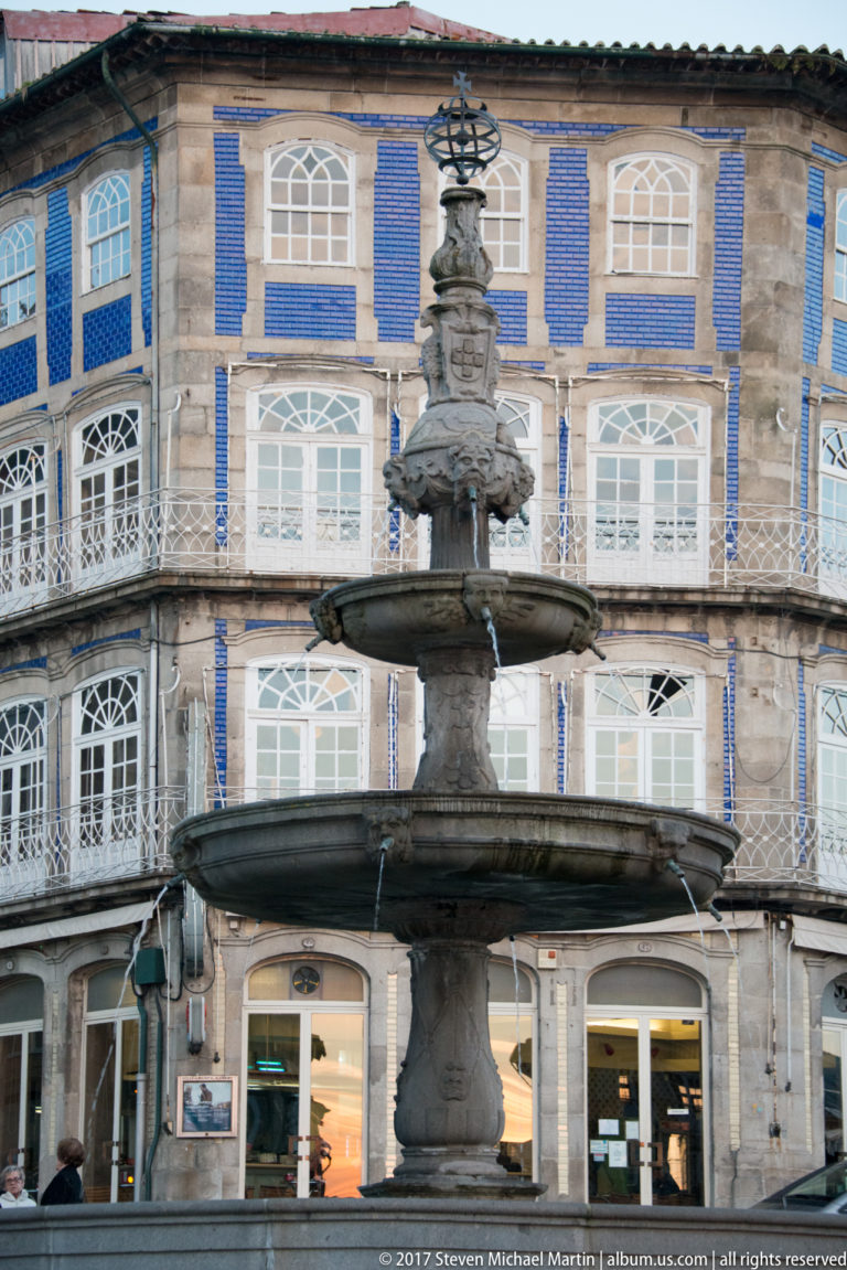 Multi-tiered stone fountain before a curved blue-and-white historic façade with rows of windows and ground-floor shops.
