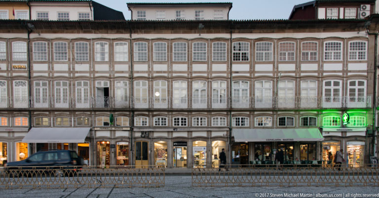 Long historic façade with dense rows of white windows, brown framing, ground-floor shops, arcades, and evening lights.