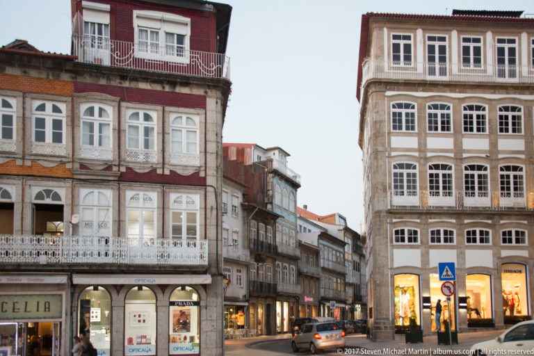 Historic corner buildings with enclosed balconies, white windows, granite trim, shops, and a narrow street leading away at dusk.