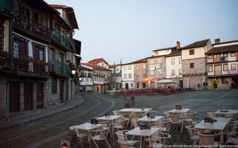 Empty white café chairs and tables in a historic Guimarães square at dusk, surrounded by traditional façades and warm lights.