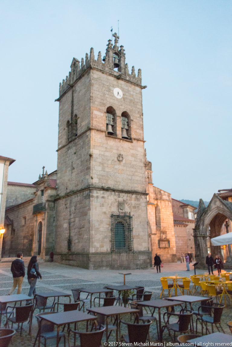 Granite Church of Nossa Senhora da Oliveira with crenellated bell tower across a café-filled square at dusk.