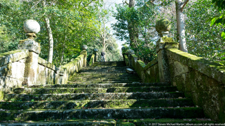 Steep mossy stone staircase rising between heavy balustrades topped by repeated spherical finials beneath trees.