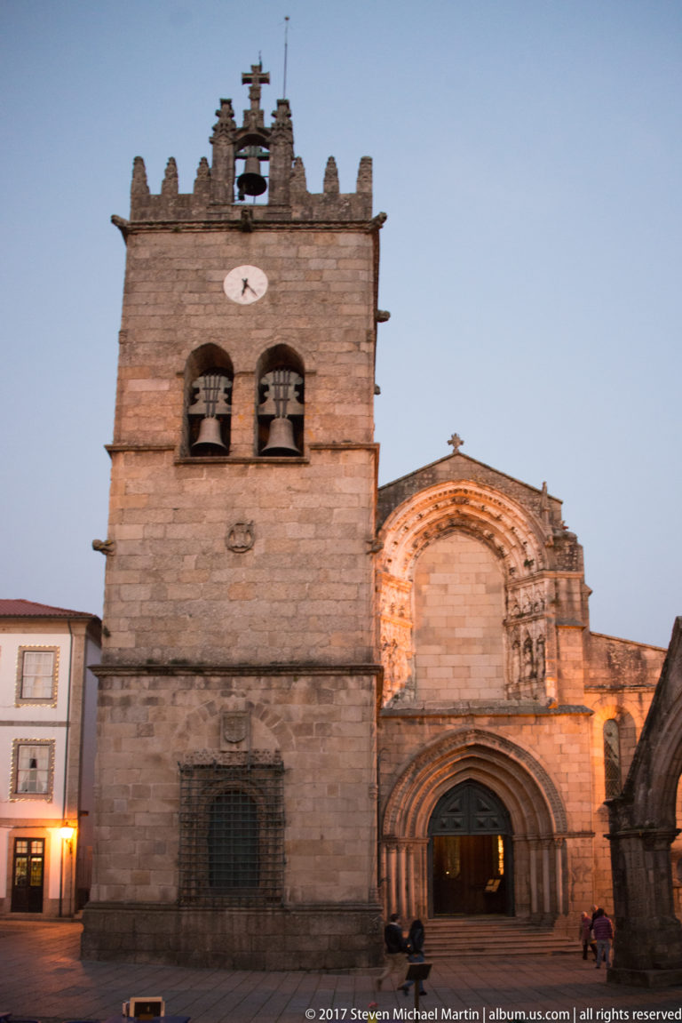 Illuminated granite church façade with deeply recessed Gothic portal, square bell tower, two visible bells, and crenellated crown at dusk.