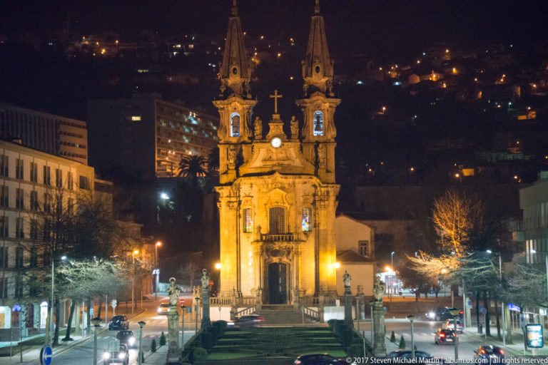 Illuminated twin-spired Nossa Senhora da Consolação e dos Santos Passos church at night, with formal garden and hillside lights behind.