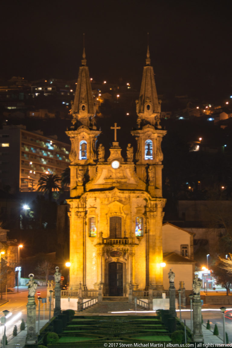Centered illuminated Baroque church with twin spires, central clock, cross, formal garden, and hillside lights at night.