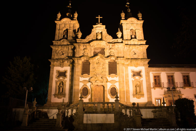 Floodlit ornate church with twin bell towers, saint niches, carved stone façade, central cross, and balustraded steps against black sky.