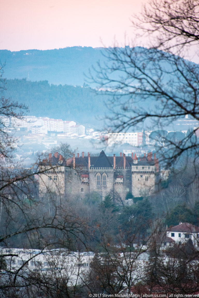 Palace of the Dukes of Braganza seen through leafless branches, with crenellated walls, tall chimneys, city buildings, and hills behind.