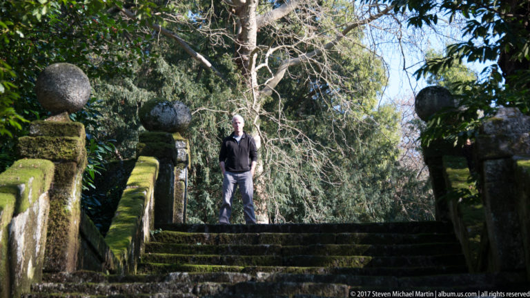 Man standing at the top of a mossy stone garden staircase between balustrades with spherical finials and tall trees.