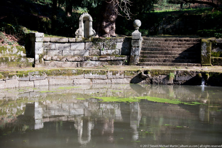 Still garden pond reflecting a stone retaining wall with arched niche, steps, ball finials, and a small water cascade.
