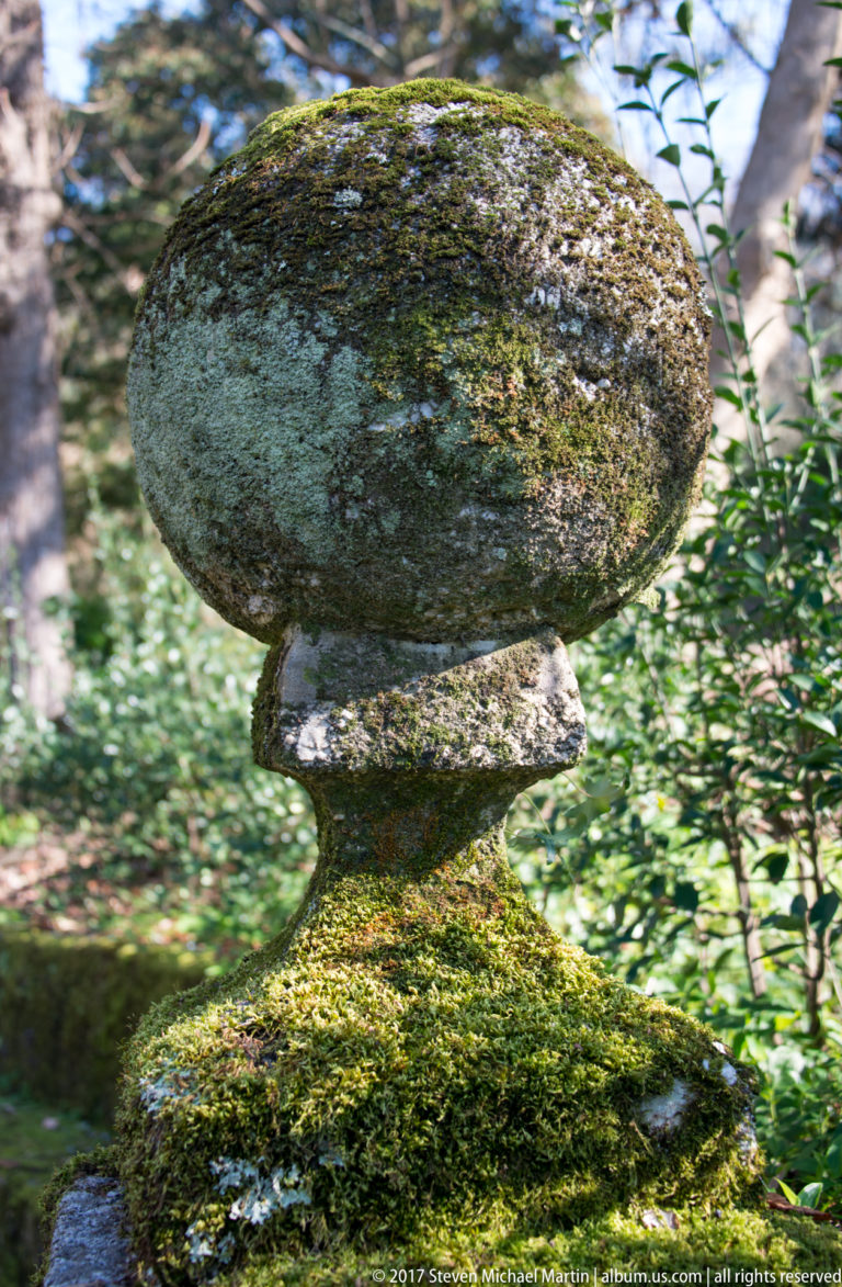 Close-up of a weathered spherical stone finial on a molded pedestal, heavily mottled with moss and lichen.