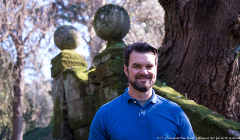 Portrait of a bearded man in a blue shirt beside a moss-covered stone balustrade with large spherical finials.