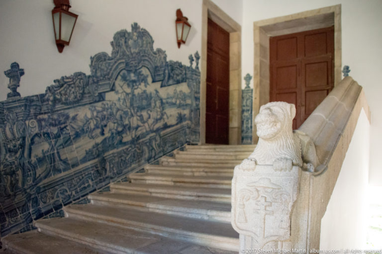 Interior stone staircase beside a blue-and-white azulejo mural, carved stone newel, and heavy stone-framed wooden doors.
