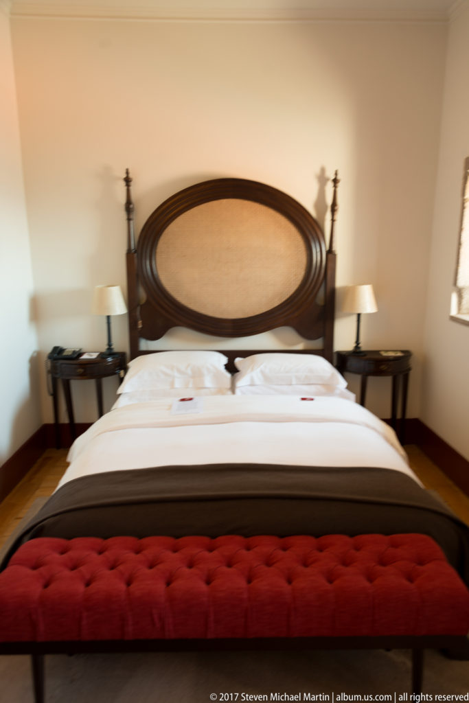 Guest room with large bed, carved dark-wood headboard, matching lamps, bedside tables, and a deep red upholstered bench.