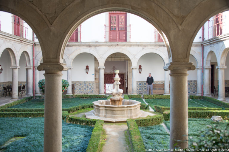 Cloister seen through rounded stone arches, with central fountain, geometric hedges, arcades, columns, and blue-and-white tilework.