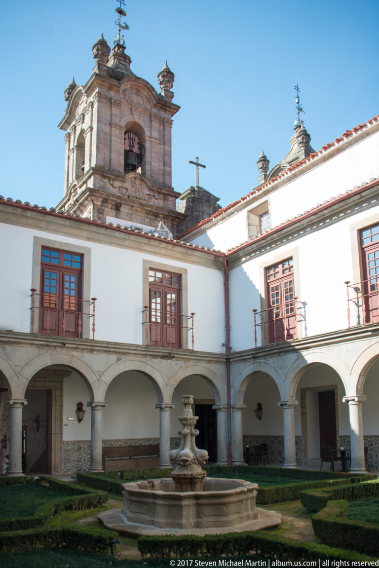 Whitewashed cloister courtyard with arcades, formal hedges, central stone fountain, red-framed windows, and church bell tower above.