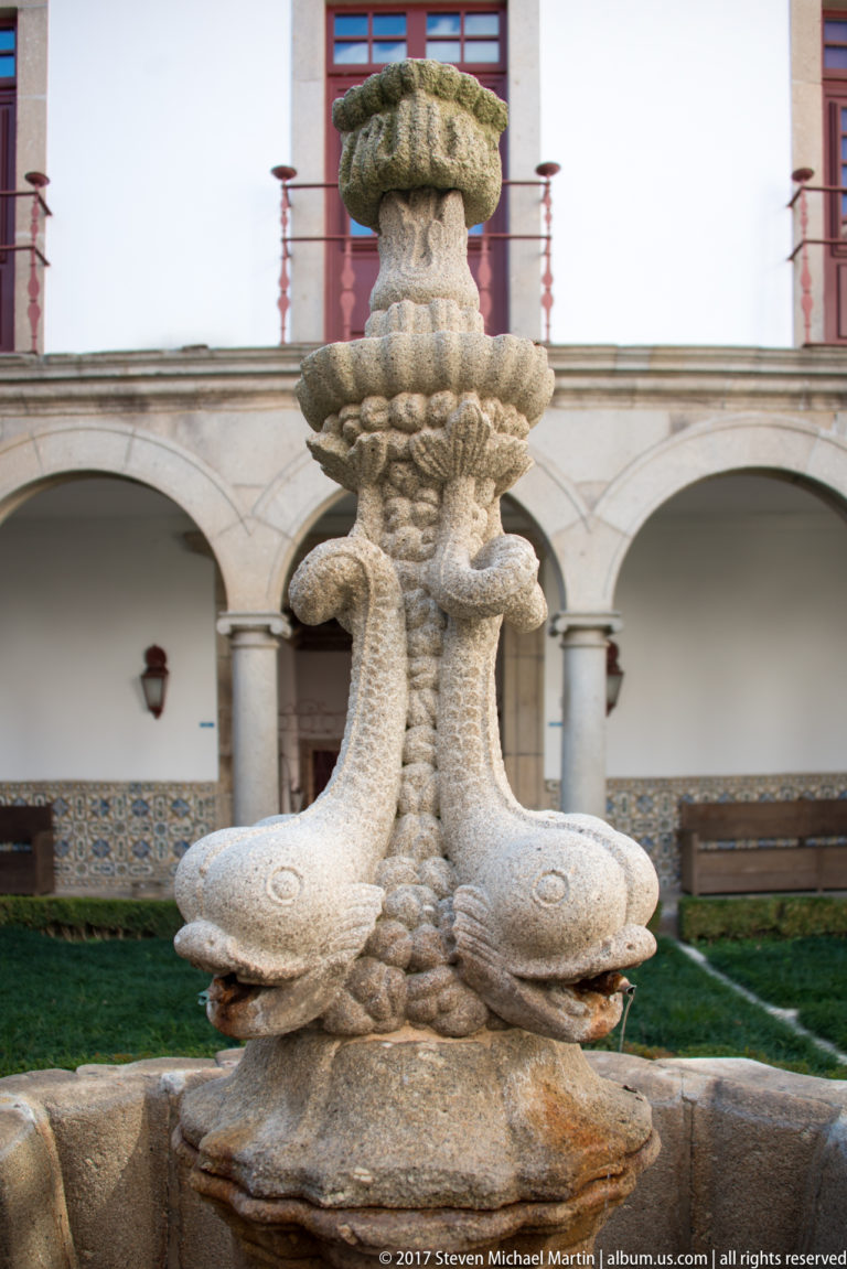 Close view of an ornate pale-stone fountain centerpiece with four projecting sculpted forms and a carved central shaft.