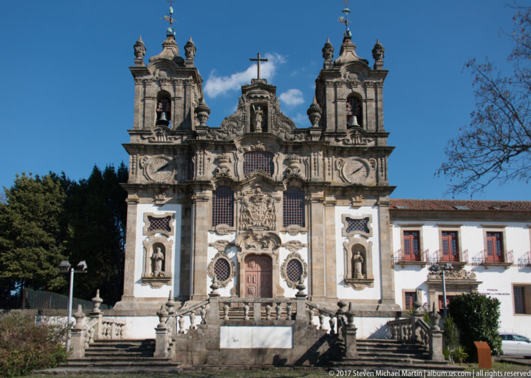 Full frontal view of an ornate white church with twin bell towers, sculpted portal, niches, circular windows, and broad stone steps.