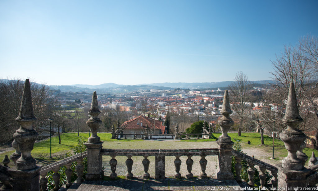 Stone terrace balustrade with decorative finials overlooking green gardens, the city of Guimarães, and distant hills under blue sky.