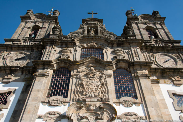 Low-angle close view of ornate church upper façade with twin towers, clocks, barred windows, heraldic carving, sculpted figures, and cross.