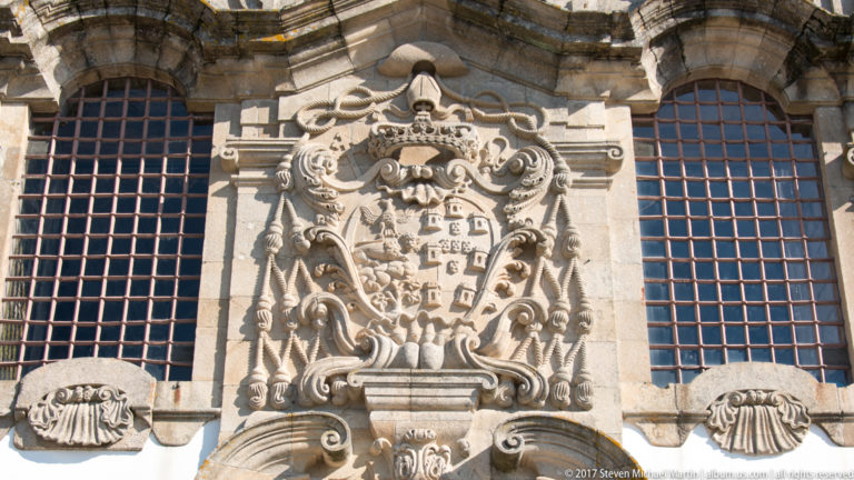 Close view of a crowned stone coat of arms surrounded by scrollwork and sculpted figures between tall barred church windows.