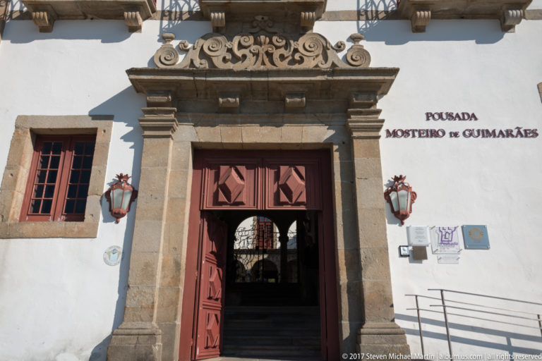 Entrance marked “Pousada Mosteiro de Guimarães,” with red wooden doors, granite surround, wall lanterns, plaques, and cloister visible inside.
