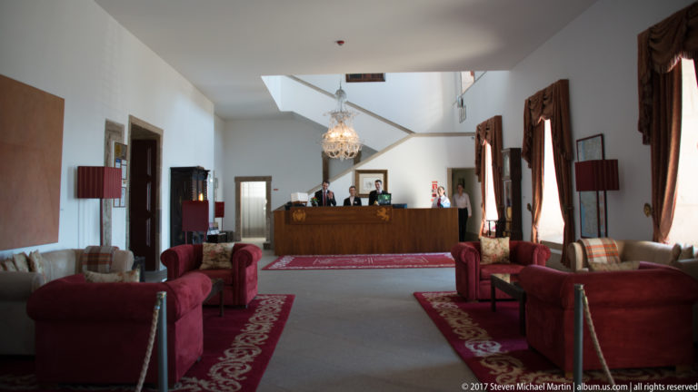 Spacious hotel reception lounge with wooden desk, chandelier, red armchairs, sofas, table lamps, and patterned carpets.