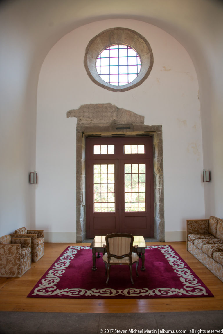 White-walled sitting room with oval oculus, stone-framed glazed doors, patterned sofas, red carpet, and a small central table.