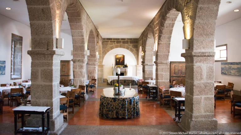 Dining room with broad granite arches and piers, white-clothed tables, wooden chairs, central table, and warm wall lighting.