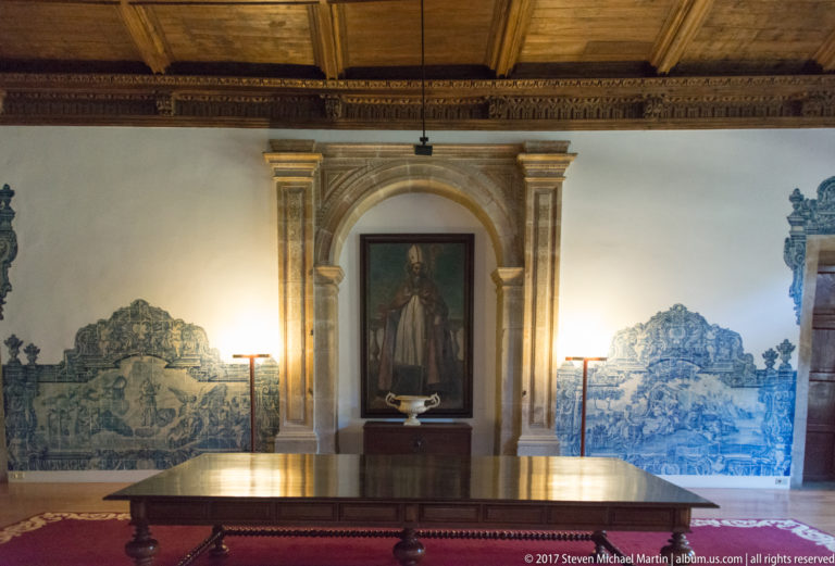 Formal room with blue-and-white azulejo panels, a stone-framed religious portrait, long wooden table, and decorated timber ceiling.