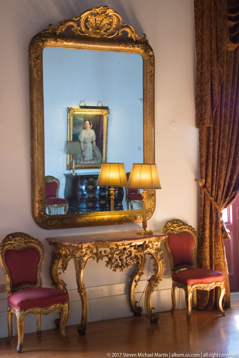 Large gilt mirror above an ornate console table, flanked by red upholstered chairs and lamps beside heavy patterned curtains.