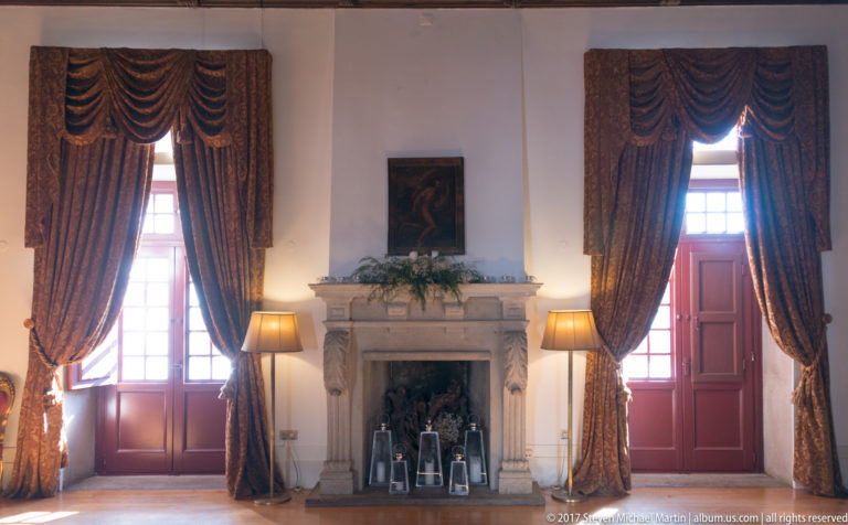 Stone fireplace centered between two tall curtained windows, with paired lamps and small framed objects arranged in the hearth.
