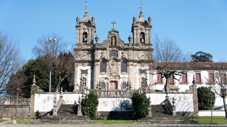 Ornate twin-towered church with granite carving, central cross, broad stairways, and a walled forecourt beneath a clear blue sky.