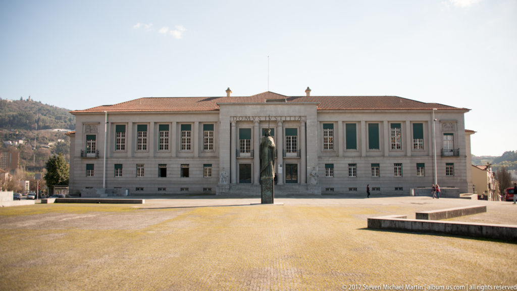Symmetrical pale-stone civic building with tall windows and central entrance behind a freestanding monument in a broad paved square.