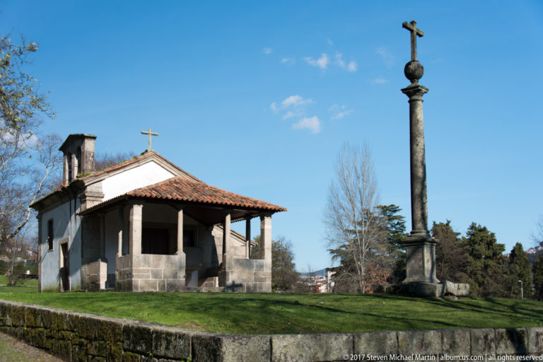 Small white chapel with tiled roof and open porch beside a tall stone column topped by a cross on a grassy rise.