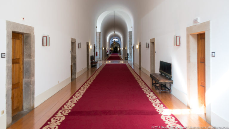 Long white vaulted corridor with red carpet runner, stone-framed wooden doors, benches, framed art, and repeating arches.