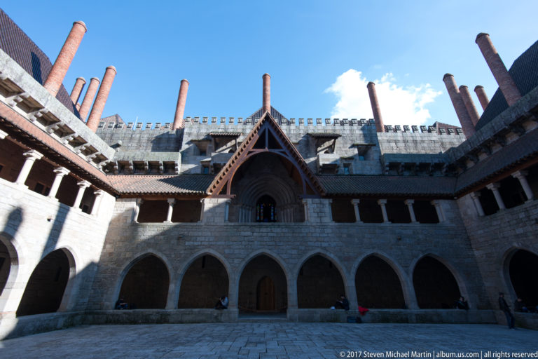 Stone palace courtyard with two-level arcades, steep roofs, tall brick chimneys, and a projecting gabled upper opening.