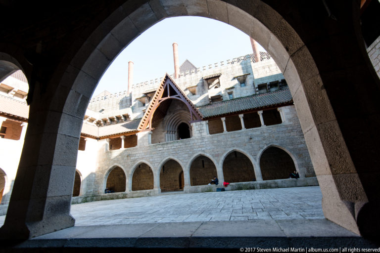 Sunlit palace courtyard and arcades seen through a dark granite arch, with steep roof, tall chimneys, and projecting gable.