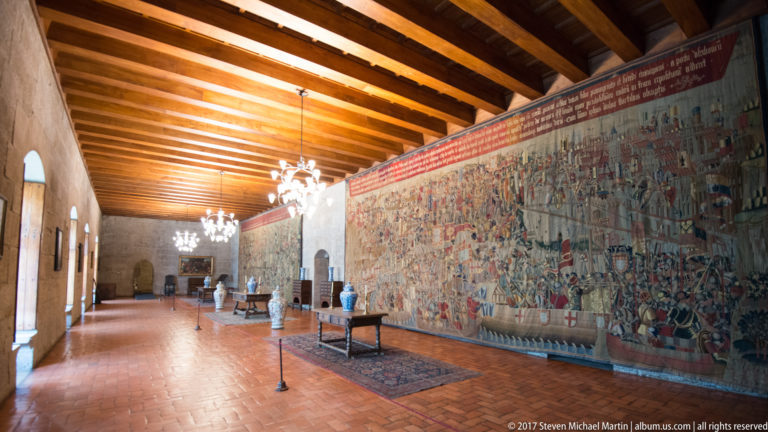 Long stone hall with exposed timber ceiling, chandeliers, furniture, and a monumental narrative tapestry covering the right wall.
