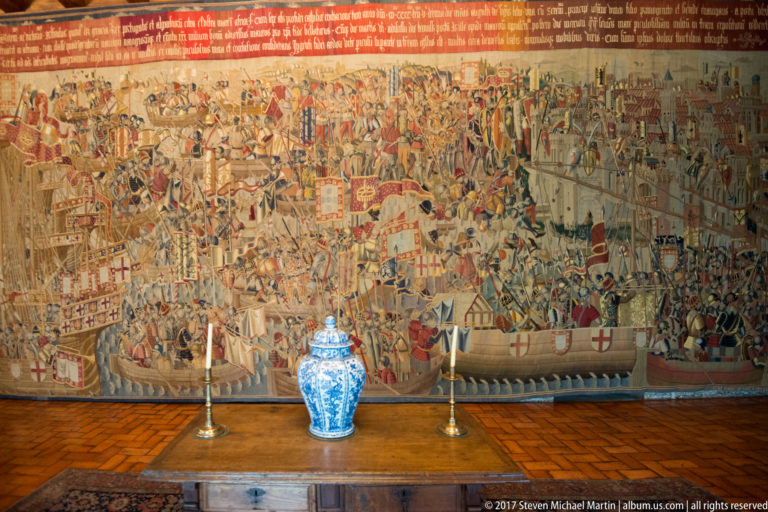 Large historic tapestry crowded with ships, soldiers, banners, and architecture above a table with blue-and-white porcelain and candlesticks.