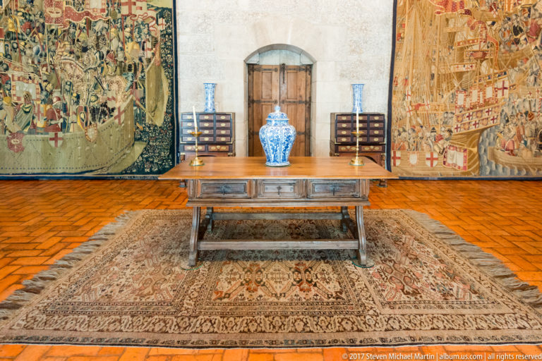 Wooden table on a patterned carpet between two tapestries, with blue-and-white porcelain vases, drawer cabinets, and a central door.
