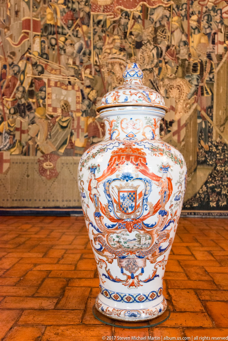Large lidded porcelain jar painted in blue, red-orange, and green with scrolling decoration, displayed before a tapestry.