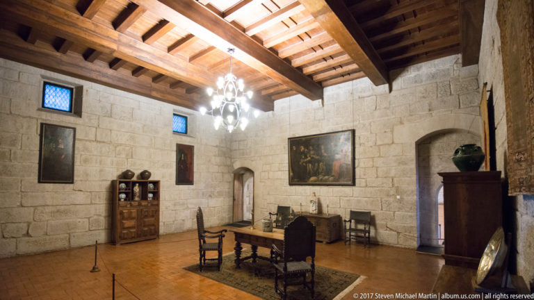 Granite palace room with exposed timber ceiling, chandelier, dark table and chairs, paintings, cabinet, and small blue-glazed windows.
