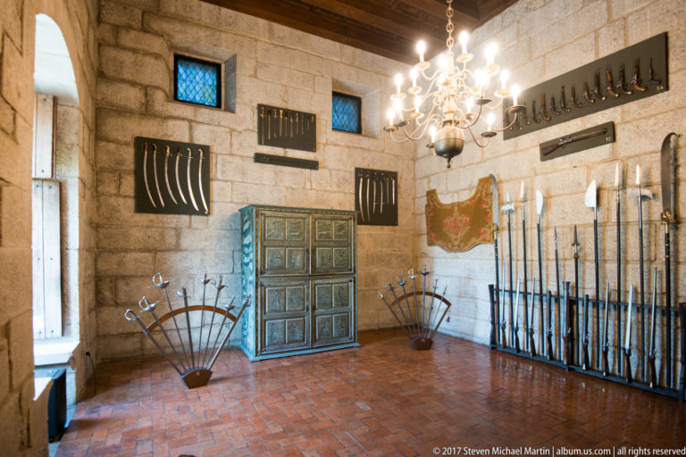 Stone museum room displaying rows of polearms, swords, curved blades, and fan-shaped weapon arrangements around a painted cabinet.