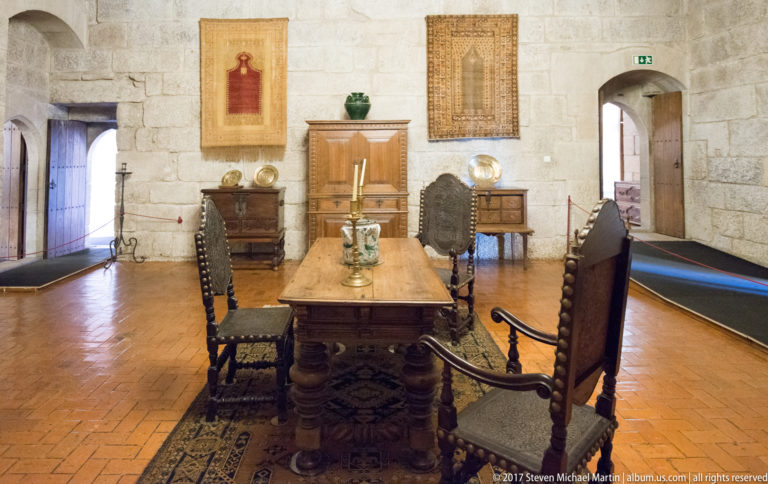 Historic dining room with heavy wooden table, high-backed chairs, patterned carpet, cabinets, ceramics, and wall hangings against granite walls.