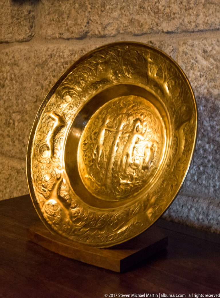 Shallow golden metal dish on a wooden stand, densely embossed with figures, foliage, and decorative bands against a stone wall.