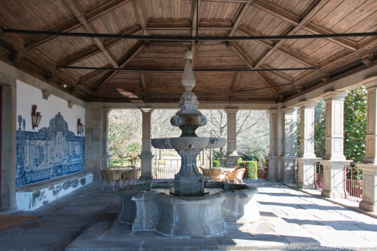 Tiered stone fountain beneath an ornate timber ceiling in an open loggia with columns and blue-and-white tile panels.