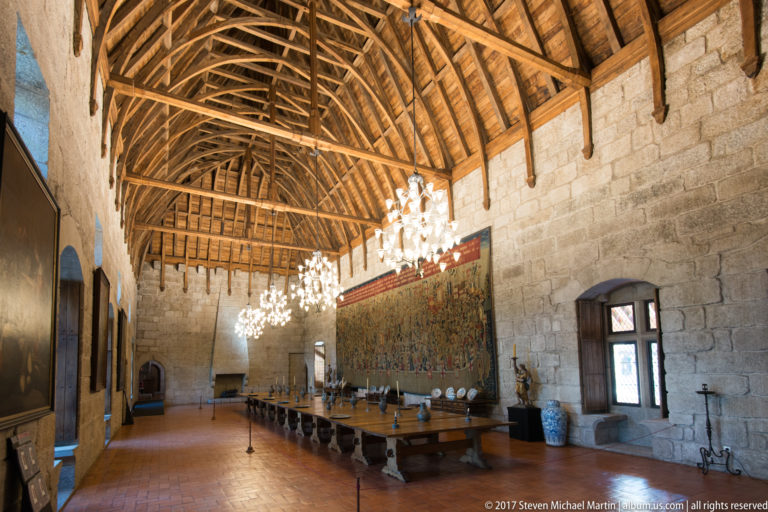 Long granite hall beneath a high timber roof, with chandeliers, extended banquet table, ceramics, and a monumental wall tapestry.