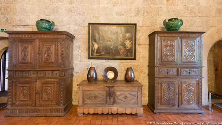Two tall carved wooden cabinets flank a lower chest topped with ceramics and a round dish beneath a framed painting.